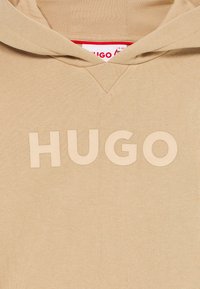 Sweatshirt bege com capuz, com letras em relevo a dizer "HUGO" no peito e uma etiqueta vermelha e branca dentro do colar.