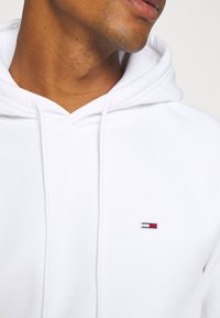 Sudadera blanca de algodón suave, con un bolsillo frontal, capucha con cordón y un pequeño logotipo bordado con acentos en rojo, blanco y azul.