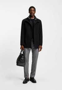 Uomo con cappotto nero, pantaloni grigi, mocassini neri, che tiene una borsa di pelle nera, in piedi contro uno sfondo bianco semplice.