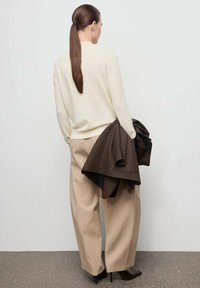 Cremefarbenes Sweater, beiger, weiter Stoffhose und ein brauner Mantel, der über dem Arm hängt. Glatte Texturen und maßgeschneiderte Designs mit einem minimalistischen ästhetischen Ansatz.