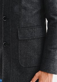 Grafitowo-szary płaszcz wełniany w jodełkę z zaokrąglonym kołnierzem, zapinany na guziki z przodu, z pojedynczą naszywaną kieszenią po lewej stronie.