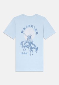 Šviesiai mėlyna medvilninė marškinėlių su cowboy ant arklio grafika, su užrašu "WRANGLER" viršuje ir "1947" apačioje tamsesnės mėlynos spalvos kontrastu.