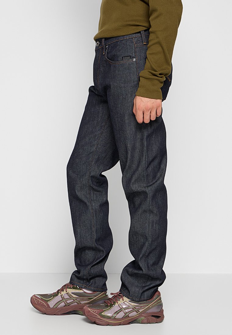 Person in dunkelblauen Jeans, olivgrünem Langarmshirt und braunen sowie olivfarbenen Sportschuhen, der vor einem weißen Hintergrund steht.