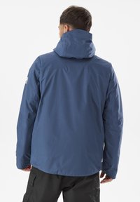 Man facing away wearing a blue hooded jacket and black cargo pants against a plain white background.