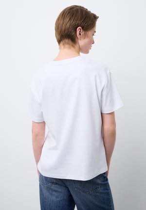 Person mit kurzen Haaren, die ein schlichtes weißes T-Shirt mit kurzen Ärmeln und eine blaue Jeans trägt, von hinten vor einem weißen Hintergrund gesehen.