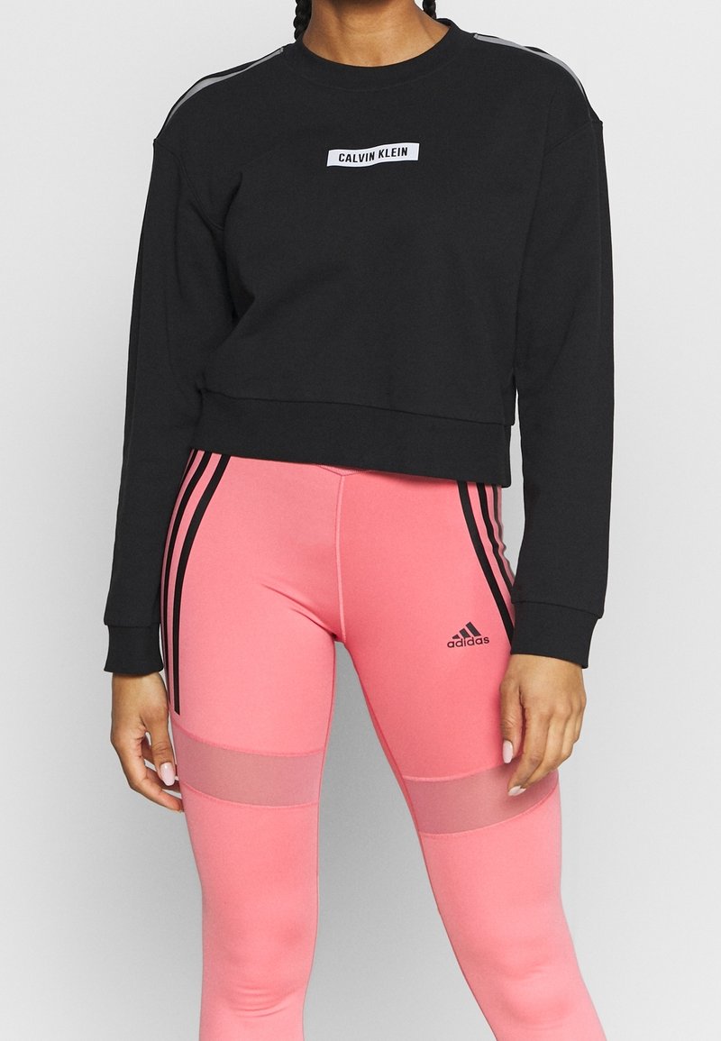 Sweatshirt noir court avec le logo "Calvin Klein", associé à des leggings roses avec des rayures noires et des panneaux en maille pour la respirabilité.