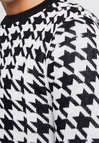 Svetr s kostkovaným vzorem houndstooth v černobílé barvě, s ribbovanou strukturou a kulatým výstřihem, vyrobený z pleteného materiálu.