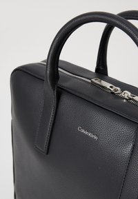 Schwarze Leder-Shopper-Tasche mit strukturierter Oberfläche, doppelte Obergriffe, silberne Beschläge und Reißverschluss. Mit einem dezenten Logo-Detail.