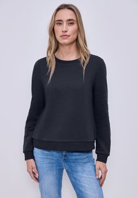Femme aux longs cheveux blonds portant un sweat-shirt noir uni à manches longues et un jean bleu clair, debout devant un fond uni.