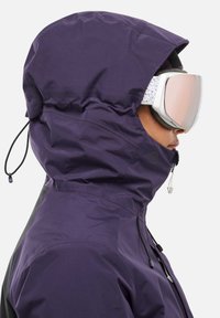 Veste imperméable violette avec une capuche ajustable. Comprend une fermeture éclair à l'avant, des cordons de serrage et une doublure intérieure à motif. Portée avec un masque de ski.