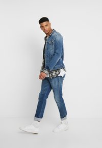 Jachetă din denim cu un finisaj albastru estompat, purtată peste o cămașă în carouri alb-negru, combinată cu blugi slim-fit albaștri și adidași albi.