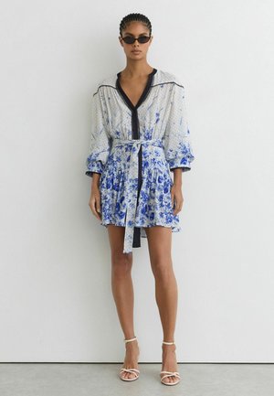 Frau trägt kurzes weißes Kleid mit blauen Blumen- und Punkten, schwarzem Abschluss, Gürtel und weißen Riemchen-Absätzen, steht vor einer weißen Wand.