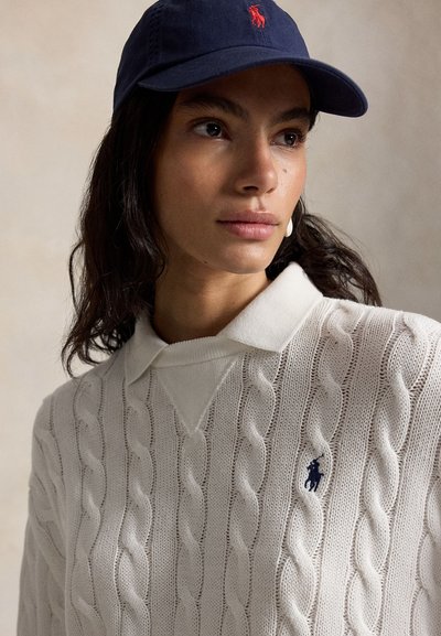 Ženska z dolgimi temnimi lasmi, ki nosi mornarsko modro kapo in belo pulover z vzorcem pletenja kabla, ovratnikom in vezenim mornarsko modrim logotipom.