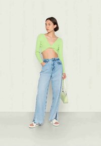 Suéter verde cropped com mangas longas, combinado com jeans de ganga azul claro de cintura alta com fendas laterais. Sandálias brancas com plataforma e uma pequena mala verde.
