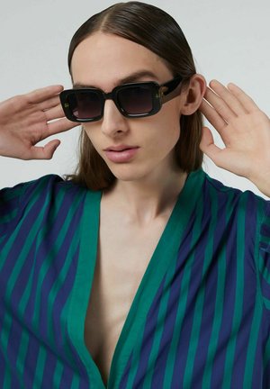 Zwarte vierkante zonnebril met verlooplenzen. Het model draagt een blauw-groen gestreepte shirt. De shirt heeft een wijde halslijn en korte mouwen.