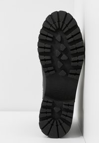 Semelle de botte en caoutchouc noir avec des crampons profonds, présentant une surface texturée et des motifs angulaires prononcés ; idéale pour la traction sur divers terrains.