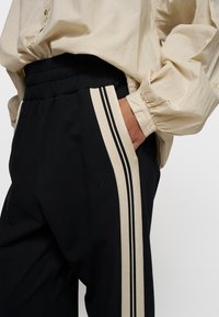 Zwarte broek met een relaxte pasvorm, voorzien van een elastische tailleband, zijvakken en beige strepen langs de zijkanten. De stof lijkt glad te zijn.