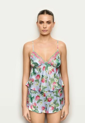 Frau trägt hellblaues Satin-Camisole und Shorts mit pinkem Erdbeer- und grünem Blättermuster, steht vor neutralem Hintergrund.