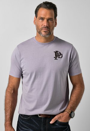 Mand med kort mørkt hår og skæg iført en lyse lilla t-shirt med sort logo på brystet, mørke jeans og sort armbåndsur, stående foran en grå baggrund.
