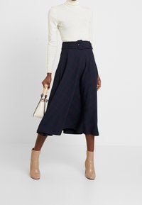 Femme portant un pull à col roulé blanc, une jupe midi trapèze bleu marine avec ceinture, des bottines beiges, et tenant un petit sac à main crème.