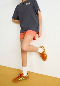 Person som bär en svart Roxy-t-shirt, orange mönstrade shorts, vita strumpor och orange sneakers med neongröna ränder, stående på ett ben.