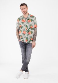 Mann mit tätowierten Armen, der ein hellbeiges Blusenshirt mit Blumenmuster, graue zerrissene Jeans und weiße Turnschuhe trägt, steht mit übereinander geschlagenen Beinen vor einem weißen Hintergrund.