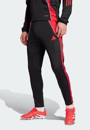 Personne portant un pantalon de sport noir à bandes rouges, un haut assorti et des chaussures de football rouges, debout sur un fond clair.