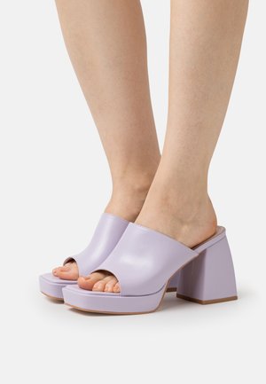 Pieds portant des mules à plateforme ouvertes en cuir lilas avec de larges bretelles et des talons carrés sur fond blanc.