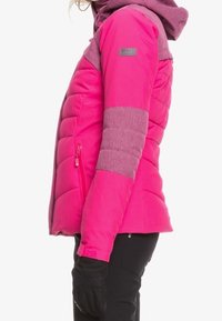 Giacca isolante rosa con cappuccio e maniche grigie testurizzate. Presenta imbottitura verticale cucita, una tasca laterale con zip e polsini regolabili.