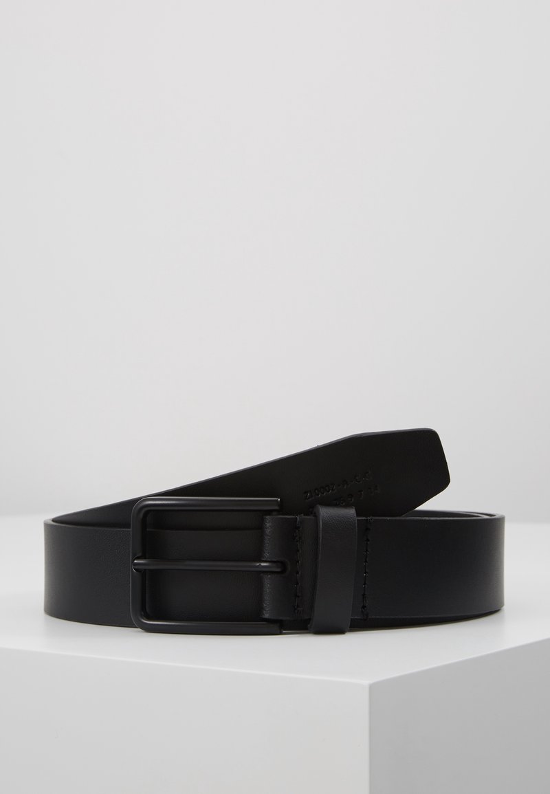 Ceinture en cuir noir avec une boucle métallique rectangulaire noire mate enroulée sur une surface blanche sur un fond gris uni.