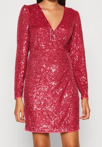 Röd paljettklänning med lång ärm, V-ringning och figurnära design. Har en jämn textur och en glänsande yta med en subtil lyster.