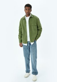 Mies seisoo käsi taskussa, päällään vihreä napitettava takki, valkoinen paita, siniset farkut ja valkoiset lenkkarit valkoista taustaa vasten.