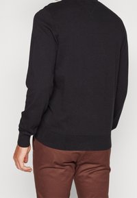 Persona che indossa un maglione lungo nero e pantaloni marroni, mostrata di spalle con la mano sinistra leggermente arricciata.