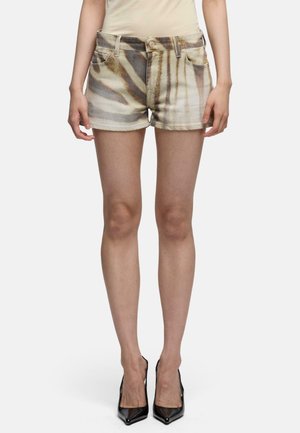 Kvinde iført beige og brun mønstret denimshorts med en beige top og sorte spidse høje hæle, stående mod en ensfarvet baggrund.