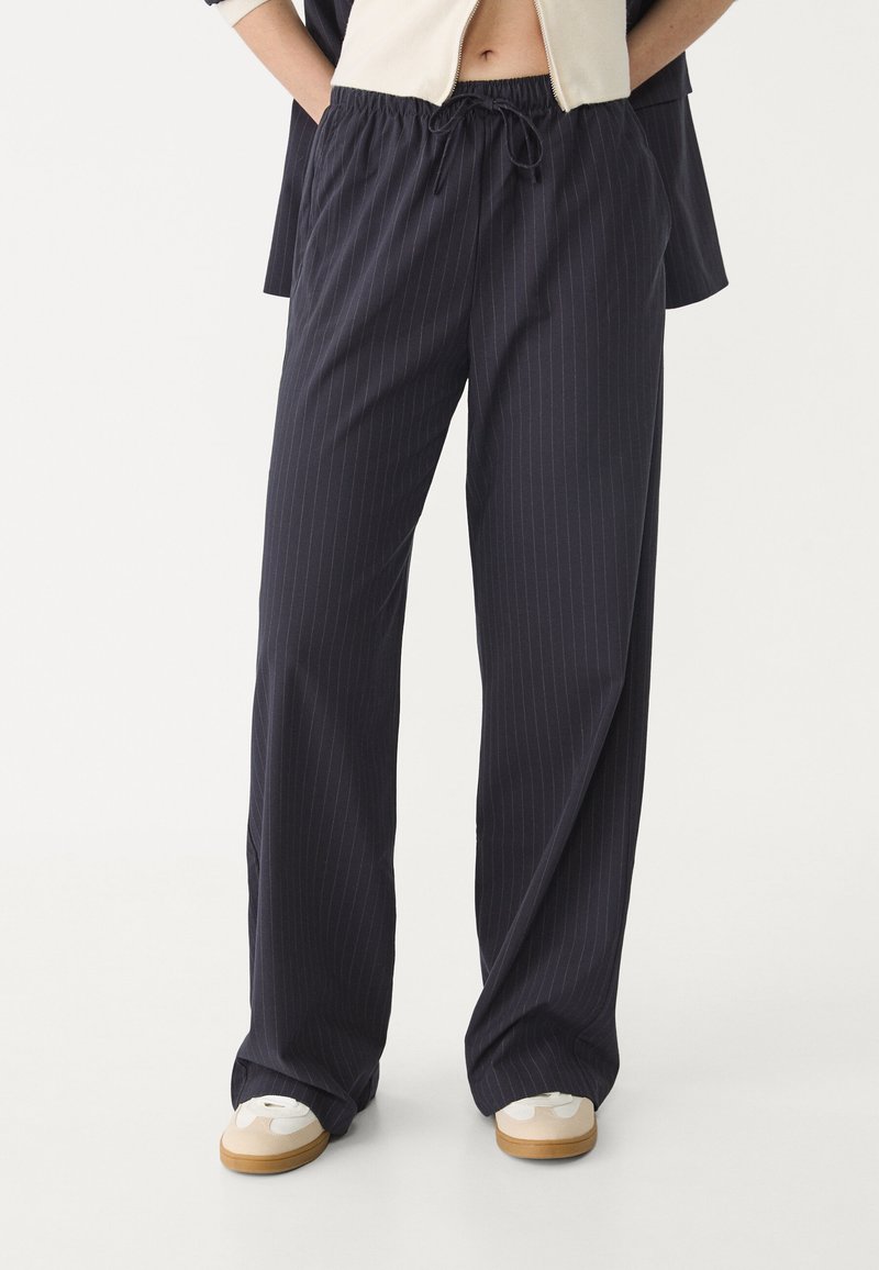 Navy gestreepte broeken met een elastische tailleband en trekkoord, losse pasvorm en zachte textuur; gecombineerd met witte sneakers.