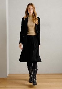 Blazer nero sopra un maglione oro textured, abbinato a una gonna nera plissettata e stivali neri fino al ginocchio. Texture lisce e design a strati.