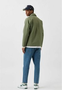 Grøn quilted jakke med krave, parret med lyseblå jeans og hvide sneakers. Hvid skjorte synlig under jakken, marineblå kasket på.