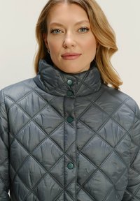 Žena so svetlými vlasmi, oblečená v tmavosivej prešívanej bunde s vysokým golierom a patentkami, hľadí priamo pred seba na neutrálnom pozadí.