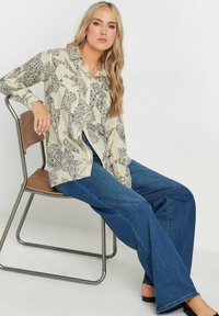 Beige shirt with black abstract pattern, buttoned, long sleeves, paired with loose blue jeans, seated on metal and wood chair.