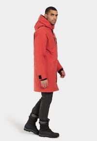 Röd vattentät parka med huva, med svarta detaljer och dragkedjestängning. Kombinerad med mörka jeans och svarta vinterstövlar.