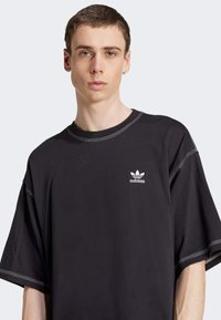 Črna oversized majica s kratkimi rokavi, izdelana iz bombaža. Opremljena z belim šivanjem in majhnim belim logotipom Adidas na prsih.