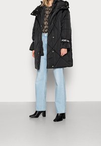 Chaqueta acolchada negra con capucha, botones a presión y detalle del logo en la manga, combinada con vaqueros de pierna recta en azul claro y botines negros.