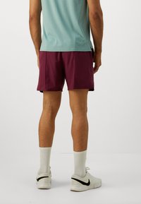 Bordeauxfarbene Sportshorts mit glatter Textur, seitlichen Taschen und lockerer Passform, kombiniert mit weißen Schuhen und Socken.