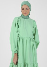 Vihreä pitkähihaista mekkoa, jossa on kerätty hame ja poimutetut yksityiskohdat. Sujuva kangas, korkea pääntie, yhdistettynä samasta kankaasta valmistettuun huiviin.