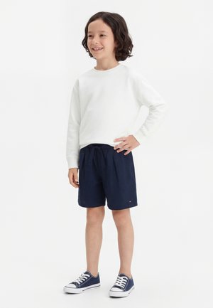 Enfant souriant aux cheveux bouclés debout, portant une chemise blanche à manches longues, un short bleu marine et des baskets bleu marine, sur un fond blanc uni.