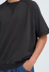 Felpa nera oversize con scollatura rotonda, maniche corte e bordo a costine. Il tessuto sembra morbido con una texture liscia e dettagli di cucitura minimi.