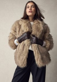 Faux fur-frakke i lys brun med en blød tekstur og bred krave; parret med brune læderhandsker og en lys sweater. Sorte bukser er synlige.