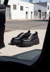 Black platform lace-up shoes placed on a sidewalk near an open car door with industrial buildings in the background.