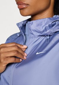 Lyseblå, letvægtsjakke med høj krave, lynlås foran og hvidt Nike-logo. Justerbare snore og strukturerede skulderdetaljer.