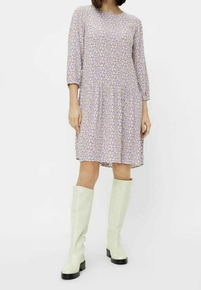Kobieta ubrana w lawendową, kwiecistą sukienkę do kolan z rękawami do łokcia oraz białe wysokie kozaki do kolan z czarnymi podeszwami.